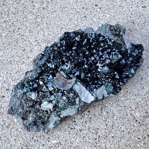 May include: A dark, irregularly shaped mineral specimen with a cluster of small, shiny, black crystals. The crystals are densely packed and reflect light, creating a sparkling effect. The base rock is a mix of gray, green, and white.