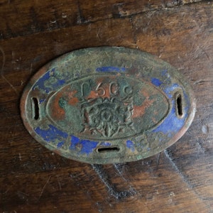 May include: A circular, oval-shaped metal tag with a blue and green enamel design. The tag has a raised design of a crest with a crown and the text "1500" in the center. The tag has two holes on the top and one hole on the bottom.