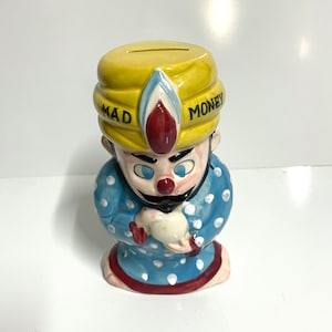 May include: A ceramic piggy bank shaped like a man wearing a yellow turban with the words "Mad Money" on it. He is wearing a blue and white polka dot robe and holding a white bird.