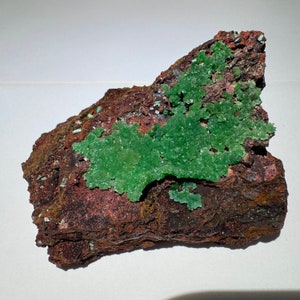 May include: A green mineral specimen with a rough, brown, and red matrix. The green mineral is clustered together in a large, irregular shape.