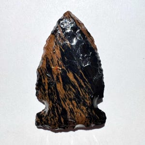 May include: A handcrafted arrowhead with a striking pattern of black and brown stripes. The stone artifact has a sharp point and a notched base, showcasing the craftsmanship of ancient tools.