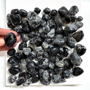 May include: A collection of numerous tumbled black stones with smooth, glossy surfaces and irregular shapes. The stones vary in size and feature natural white mineral inclusions and crystalline textures throughout the dark, polished surfaces.