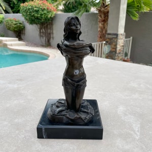 May include: Bronze sculpture of a woman holding a cloth to her chest, standing on a black marble base.
