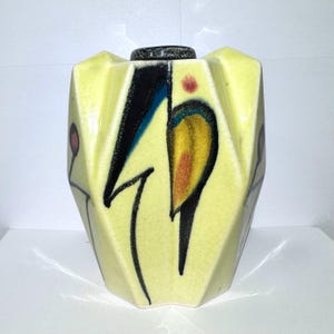May include: A pale yellow ceramic vase with a faceted, geometric body and a crackle glaze finish. The surface features abstract hand-painted designs in black, blue, and orange, including small red circular accents on the side panels.