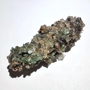 May include: A cluster of light green crystals with a rough, textured surface. The crystals are clustered together, with some areas appearing translucent. The base of the cluster has a brown and black appearance, with a few small, clear crystals.