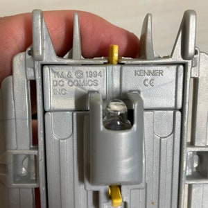 May include: Close-up of a grey plastic toy with a light bulb and a yellow switch. The toy has the text "TM & &copy; 1994 DC COMICS INC." and "KENNER CE" printed on it.