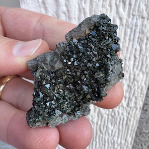May include: A dark green mineral specimen with numerous small, sparkling crystal formations. The crystals are clustered on a rough, gray matrix. The mineral has a metallic luster and reflects light, creating a shimmering effect. The overall appearance is rough and natural.