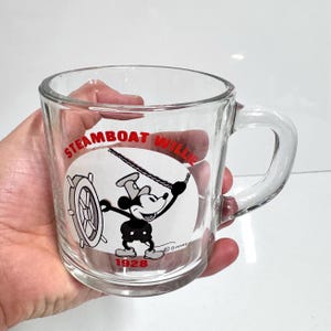 May include: A clear glass mug featuring a black and white graphic of Mickey Mouse from Steamboat Willie. The design includes the red text Steamboat Willie at the top and 1928 at the bottom, with a small Disney copyright symbol near the character.