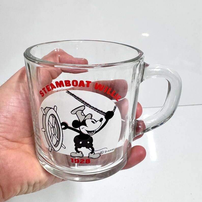 May include: A clear glass mug featuring a black and white graphic of Mickey Mouse from Steamboat Willie. The design includes the red text Steamboat Willie at the top and 1928 at the bottom, with a small Disney copyright symbol near the character.