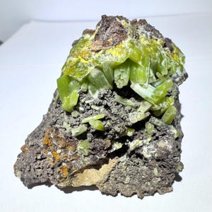 May include: A mineral specimen featuring clusters of translucent, lime-green prismatic crystals growing from a dark, porous, and textured rocky matrix. The crystalline formation displays varying shades of green with some yellow-toned highlights.