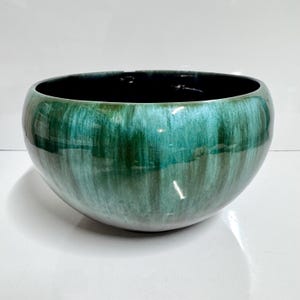 May include: A ceramic bowl with a glossy finish. The exterior is a blend of green hues, with darker shades at the top and lighter tones towards the base. The interior is a deep black. The bowl has a rounded shape.