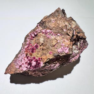 May include: A rough, irregularly shaped rock with a mix of brown, tan, and pink hues. Clusters of deep magenta crystals are visible throughout the stone. The rock has a textured surface and varying shades of color.