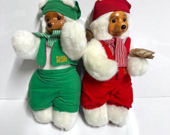Könnte beinhalten: Zwei Plüschbärenfiguren. Ein Bär trägt einen grünen Hut, Schal und Hose mit weißem Fellbesatz. Der andere Bär trägt einen roten Hut, Schal und Hose mit weißem Fellbesatz. Beide Bären haben Holzgesichter und halten Etiketten.