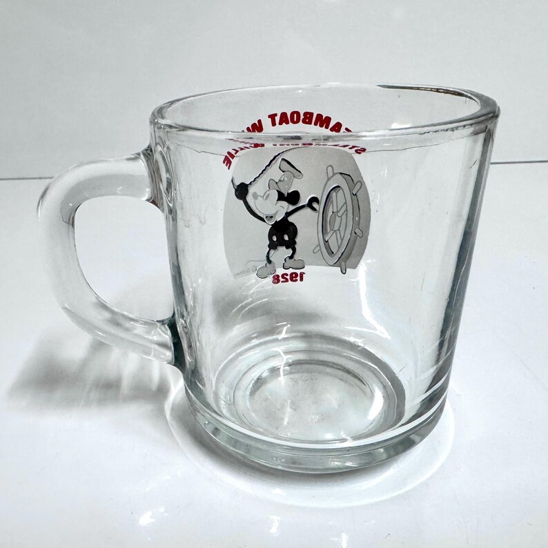 May include: A clear glass mug featuring a black and white graphic of a cartoon character at a ship's wheel. The text "STEAMBOAT WILLIE" is printed in red above the graphic, and the year "1928" is printed in red below the graphic on the glass.