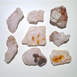 May include: A collection of nine raw, unpolished agate stones. The stones vary in shape and size, with a pale pink hue and some featuring brown and orange inclusions. The agates are displayed on a white surface.