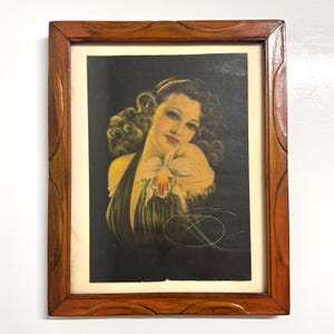 May include: Vintage framed print of a woman with dark hair and a flower. The artwork features a woman in a dark dress holding a flower, set against a black background. The frame is made of wood with a decorative edge.
