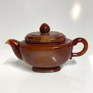 May include: A small, brown agate teapot with a lid. The teapot has a rounded body and a short spout. The lid is slightly domed and has a knob on top.