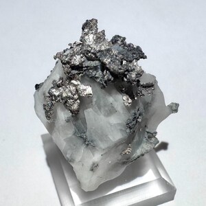May include: A raw mineral specimen featuring a cluster of metallic silver-colored native silver deposits embedded within a translucent, white crystalline quartz matrix, mounted on a clear square acrylic base for display purposes.