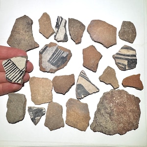 May include: A collection of ancient pottery shards in various shapes and sizes. Some shards feature black and white geometric designs, while others are plain. The color palette includes shades of brown, tan, and gray, suggesting an archaeological find.