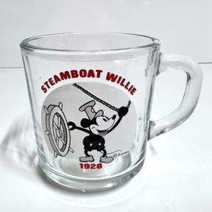 May include: A clear glass mug featuring a black and white graphic of Mickey Mouse from Steamboat Willie. The design includes the red text STEAMBOAT WILLIE at the top, the year 1928 in red at the bottom, and a small Disney copyright symbol near the character.