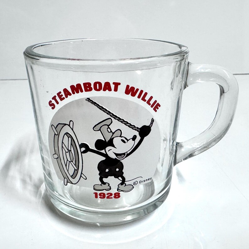 May include: A clear glass mug featuring a black and white graphic of Mickey Mouse from Steamboat Willie. The design includes the red text STEAMBOAT WILLIE at the top, the year 1928 in red at the bottom, and a small Disney copyright symbol near the character.