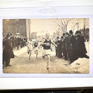 May include: Vintage sepia-toned photograph of a track and field race. Three athletes in athletic uniforms are running on a track, with spectators lining the sides. The athletes are wearing tank tops and shorts. The photograph is framed.