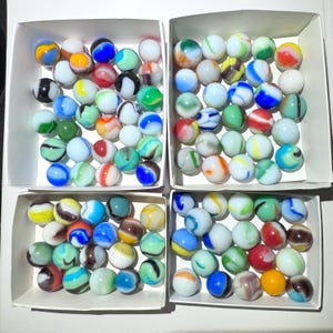 May include: Four white cardboard boxes filled with a collection of assorted glass marbles. Each marble features unique swirling patterns, opaque and translucent finishes, and a variety of colors including blue, green, red, yellow, and white.