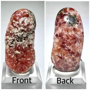 May include: A red and white crystal with a silvery gray patch on the back. The crystal is on a white stand. Front and back views of the crystal are shown.