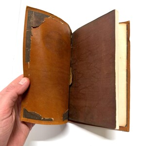 K&ouml;nnte beinhalten: Ein braunes, ledergebundenes Buch mit einem abgenutzten Einband und ein paar offenen Seiten. Das Buch wird von einer Hand offen gehalten.