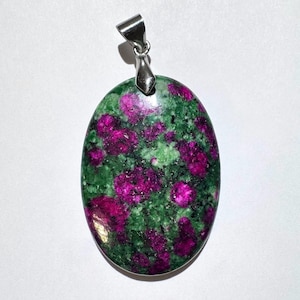 May include: Oval-shaped pendant with a vibrant green and magenta stone. The stone has a speckled pattern, with magenta spots scattered across a green base. A silver-colored bail is attached at the top for hanging.