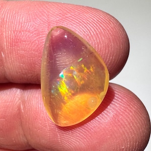 May include: A polished, teardrop-shaped opal gemstone. The opal displays a vibrant play-of-color with flashes of green, yellow, and orange. The stone is held between fingers, showcasing its translucent quality and internal patterns. The opal's surface is smooth and reflective.