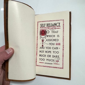 K&ouml;nnte beinhalten: Ein ge&ouml;ffnetes Buch mit braunem Lederumschlag. Die Seite ist cremefarben mit rotem und schwarzem Text. Der Text lautet "SELF RELIANCE DO THAT WHICH IS ASSIGNED YOU AND YOU CAN - NOT HOPE TOO MUCH OR DARE TOO MUCH EMERSON".