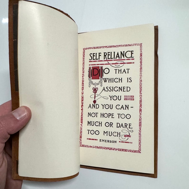 K&ouml;nnte beinhalten: Ein ge&ouml;ffnetes Buch mit braunem Lederumschlag. Die Seite ist cremefarben mit rotem und schwarzem Text. Der Text lautet "SELF RELIANCE DO THAT WHICH IS ASSIGNED YOU AND YOU CAN - NOT HOPE TOO MUCH OR DARE TOO MUCH EMERSON".