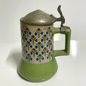 May include: A green and white ceramic beer stein with a pewter lid and a geometric pattern of red, blue, and green stylized flowers. The stein has a handle and a spout.