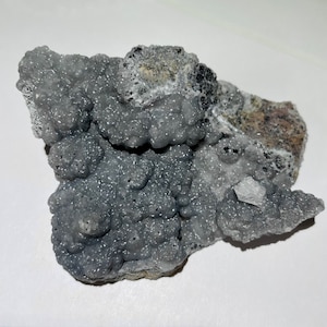 May include: A cluster of gray, sparkly, botryoidal smithsonite crystals on a matrix of dark gray rock. The crystals have a rough, textured surface and are arranged in a random pattern.
