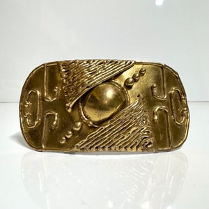 May include: A gold-toned metal belt buckle with an abstract design featuring a central circle and geometric shapes.