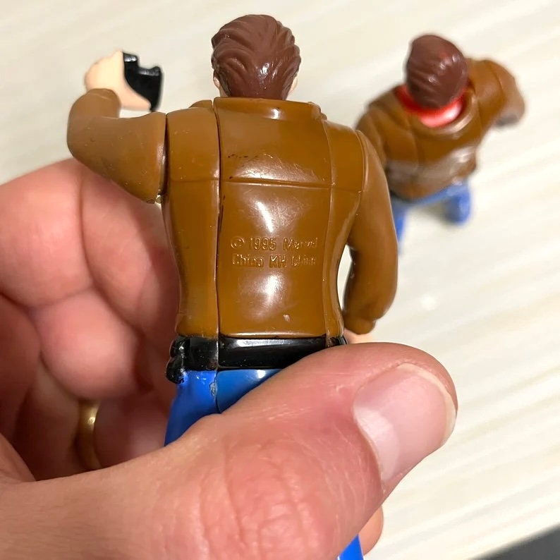 May include: Two brown plastic action figures wearing blue jeans. The figure on the left is holding a black gun. The figure on the right is facing forward. The figure on the left has the text "&copy;1995 Marvel  Disney  KH  Ltd" on the back.
