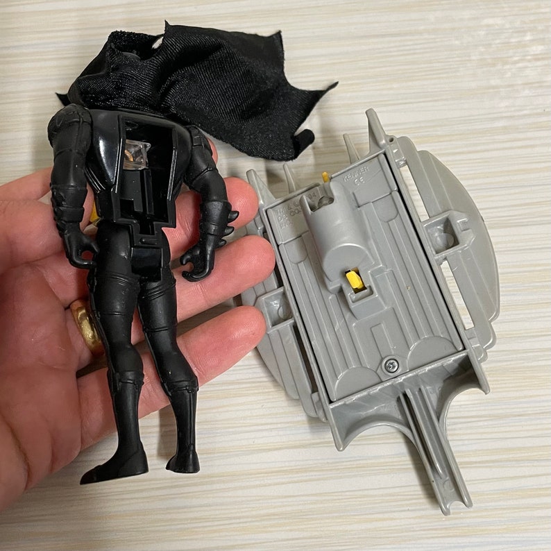 May include: Black plastic action figure of Batman with a black cape. The figure is standing on a grey plastic base with a yellow button. The base is shaped like a batarang.