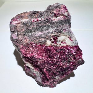 May include: A rough-hewn rock formation with layers of deep magenta, gray, and white. The magenta layer has a crystalline appearance. The rock's surface is textured, showcasing the natural mineral composition. The overall aesthetic is raw and geological.