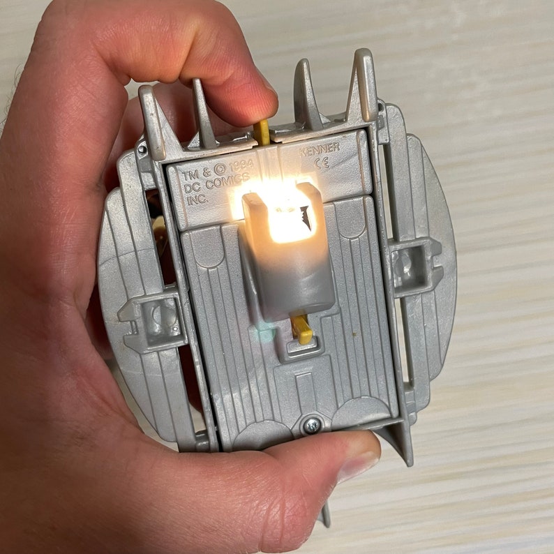 May include: A silver toy with a light-up feature. The toy has a yellow switch and the text "TM & &copy; 1994 DC COMICS INC. KENNER CE" printed on it.