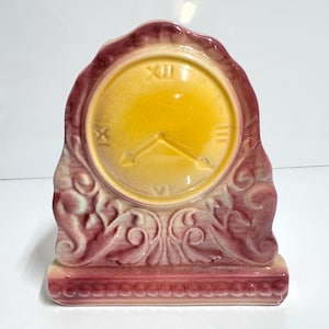 May include: A vintage ceramic clock in shades of yellow and burgundy. The clock face is yellow with black Roman numerals and hands. The clock has a decorative burgundy frame with ornate scrollwork and a rectangular base.