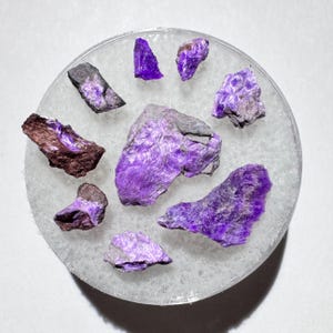 May include: A collection of raw, natural charoite gemstones in various shapes and sizes. The stones display a vibrant purple hue with swirling patterns and are arranged on a circular, frosted glass surface.