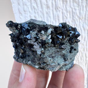 May include: A dark, rough mineral specimen featuring clusters of lustrous, black crystals. The crystals are sharply defined and sit atop a matrix of gray-green stone. The overall appearance is rugged and natural, showcasing the raw beauty of the mineral.
