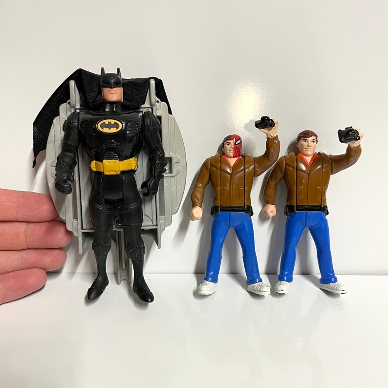 May include: Three action figures, one of Batman in a black suit with a yellow utility belt, and two other figures in brown jackets and blue jeans. The Batman figure is holding a black batarang.