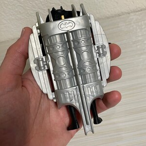 May include: A silver and black plastic toy jetpack with a Batman logo. The jetpack has two large, cylindrical engines and a black strap.
