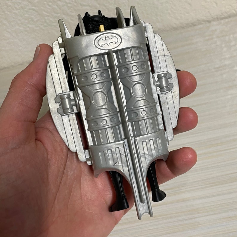 May include: A silver and black plastic toy jetpack with a Batman logo. The jetpack has two large, cylindrical engines and a black strap.
