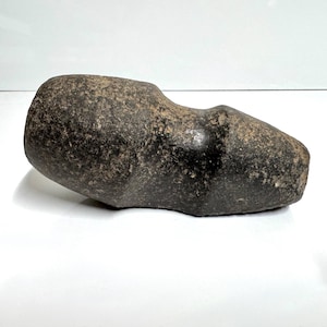 May include: A dark gray, weathered stone tool featuring a cylindrical body with a distinct central groove encircling the midsection. The surface of the stone is textured with natural mineral variations, showing a blunt, rounded end and a tapered end.