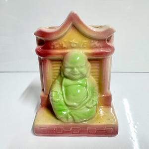 May include: A ceramic Buddha figurine in a temple-like structure. The Buddha is a vibrant green, seated in a meditative pose. The temple is a mix of red, yellow, and beige, with a small opening at the top.