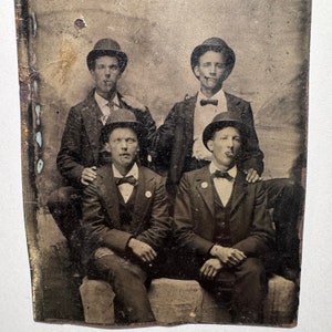 May include: A vintage black and white photograph of four men wearing hats and suits. The men are posed in a formal setting, with their hands clasped in front of them. The image is slightly faded and has a textured background.
