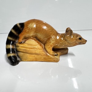 May include: A ceramic raccoon figurine in shades of brown, tan, and black. The raccoon is posed on a light brown base, with its striped tail curved upwards. The figurine has a glossy finish and is set against a white background.
